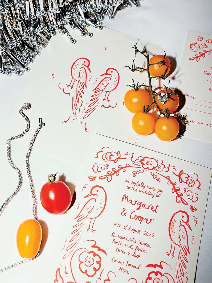 Wedding invitations with red bird and floral illustrations, featuring the names Margaret and Cooper, dated 16th of August, 2025, at St. Leonard's Church, North End, Boston, at three o'clock with summer formal attire. The invitations are accompanied by a silver chain necklace, a diamond ring placed on a cluster of yellow cherry tomatoes, and a single red cherry tomato. Silver pins are scattered in the top left corner.