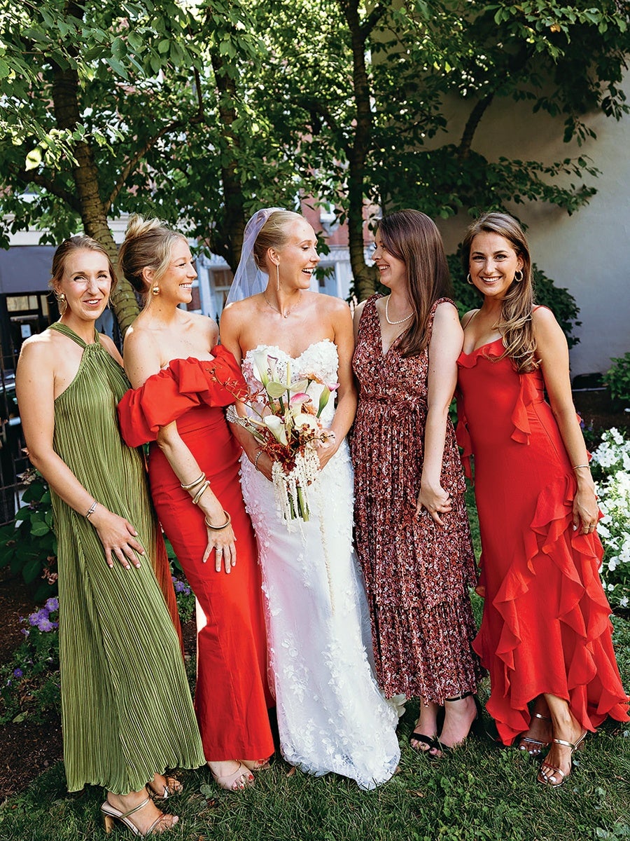 Five women stand outdoors on grass with trees and flowers in the background. The woman in the center wears a white strapless wedding gown with floral details and a veil, holding a bouquet of flowers. To her left, one woman wears a red off-the-shoulder dress with ruffles, and another wears a green halter-neck pleated dress. To her right, one woman wears a sleeveless floral dress with a pearl necklace, and another wears a red sleeveless dress with ruffles and metallic sandals. All are smiling and appear joyful.