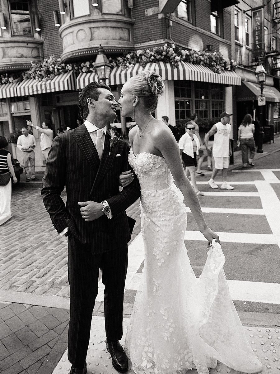 A bride and groom stand closely together on a city street, about to kiss. The bride wears a strapless, floral lace wedding gown and holds the train in one hand. The groom is dressed in a dark pinstripe suit with a tie and a boutonniere. Behind them, a building with striped awnings and flower decorations is visible, along with several casually dressed pedestrians crossing the street. The photo is in black and white.