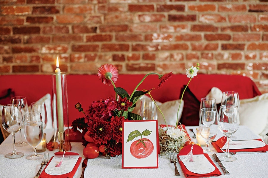 A dining table set for four with white tablecloth, red napkins, and menus. Each place setting includes a wine glass, water glass, and silverware. The centerpiece features a bouquet of red and white flowers, a tall lit candle in a glass holder, and a table number card with a red apple illustration and the number 2. The background shows a red cushioned bench and a brick wall.