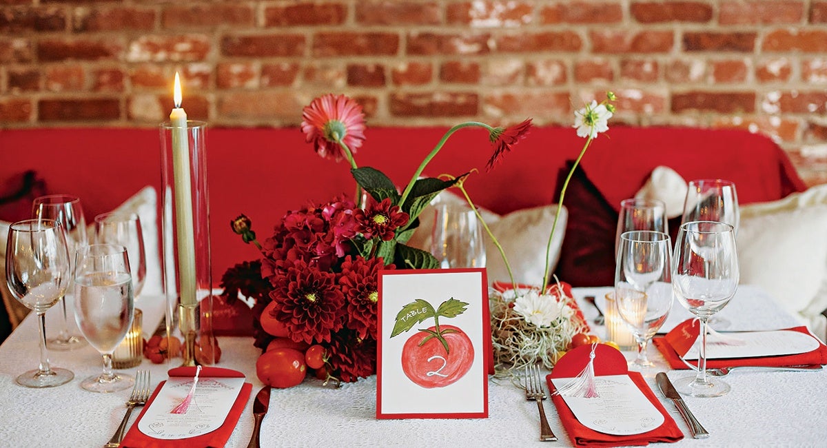 A dining table set with red napkins, white menus, and clear wine and water glasses. In the center, there is a floral arrangement with red and white flowers, including dahlias and hydrangeas, along with some tomatoes. A tall lit candle in a glass holder is on the left side. A table card with a red apple illustration and the number "2" is placed in the middle. The background features a red cushioned bench and a brick wall.