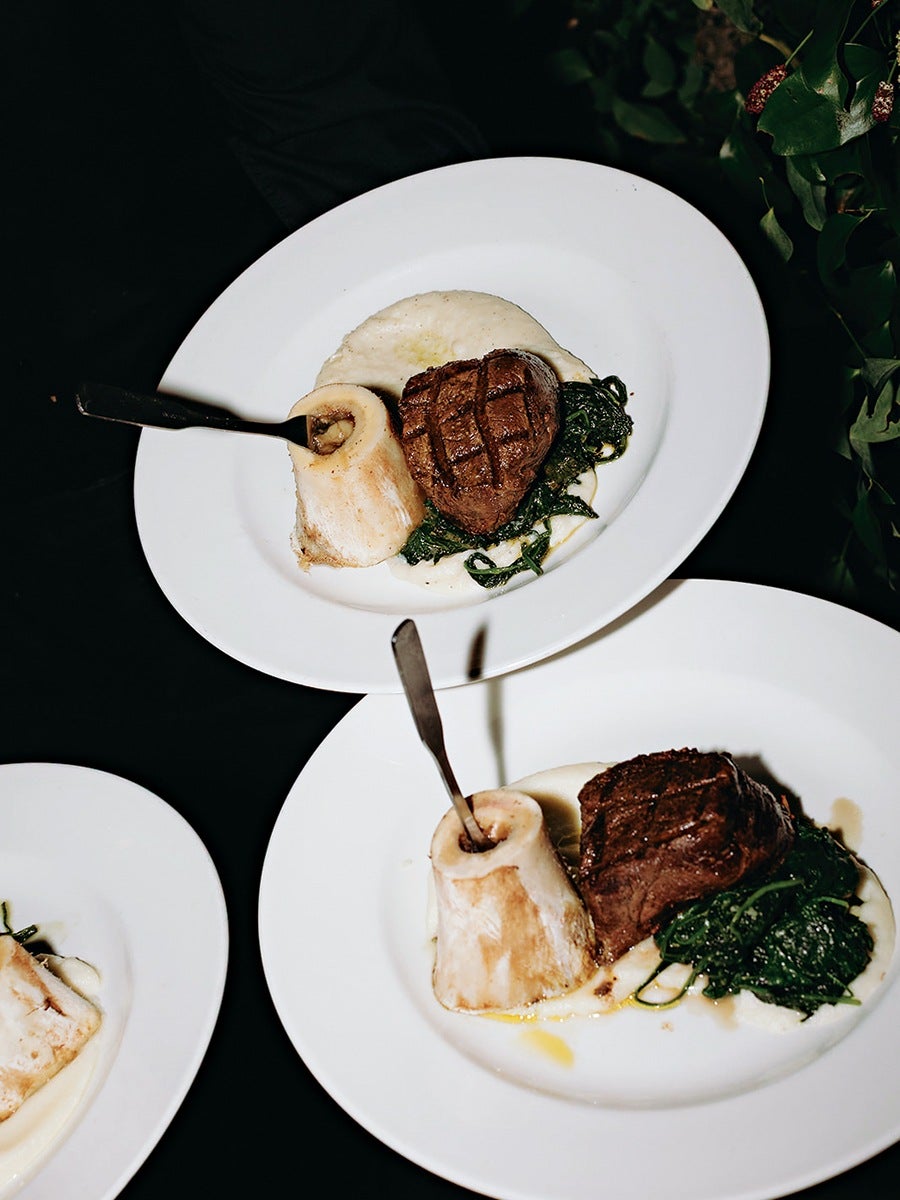 Two plates each with a grilled steak, sautéed spinach, a bone marrow piece with a spoon inserted, and a creamy white puree, all arranged on white round plates against a dark background. A third plate with bone marrow and puree is partially visible.