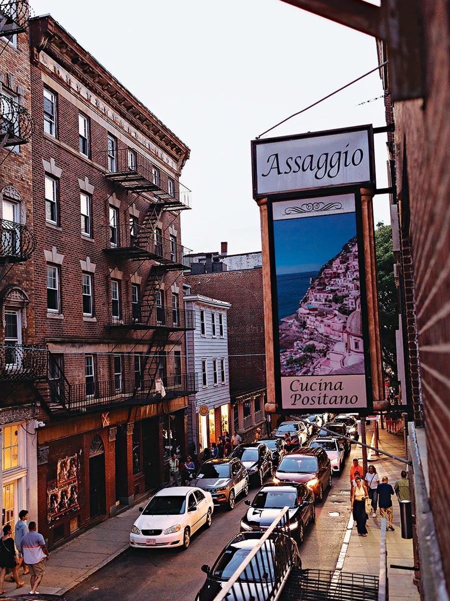 A street scene at dusk with a traffic jam of cars and pedestrians walking on the sidewalk. On the right side, a large vertical sign for "Assaggio Cucina Positano" features an image of a coastal town with colorful buildings on a hillside. The left side shows a brick building with fire escapes and windows. The atmosphere suggests a busy urban area.