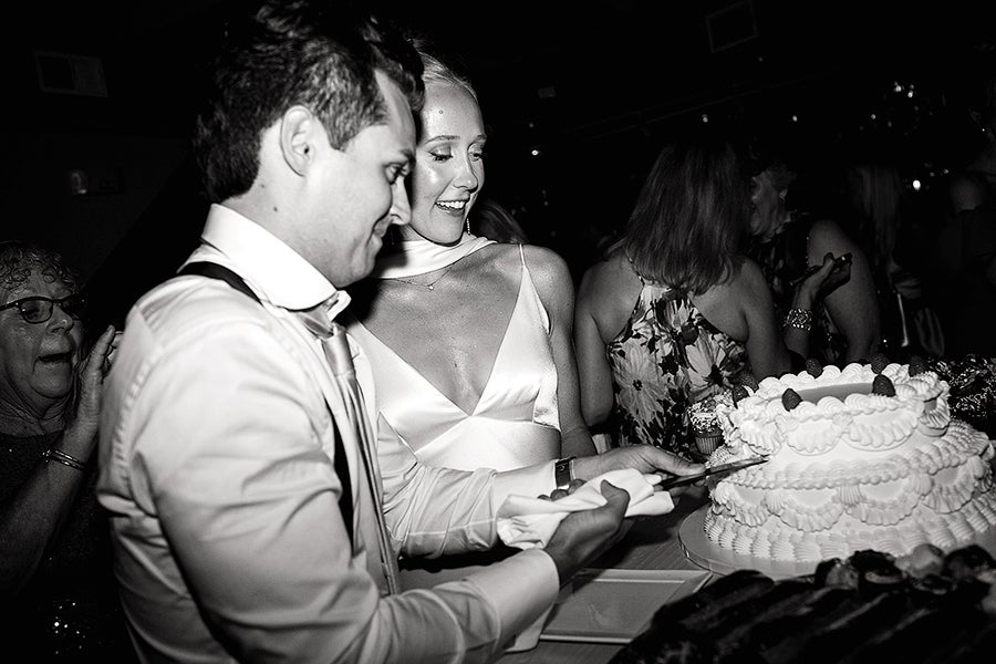 A man and woman are cutting a large, multi-layered cake decorated with piped frosting and topped with strawberries. The man is holding the knife, and the woman, wearing a satin dress with thin straps, is smiling and looking at the cake. Other people are visible in the background, including an older woman clapping. The image is in black and white.