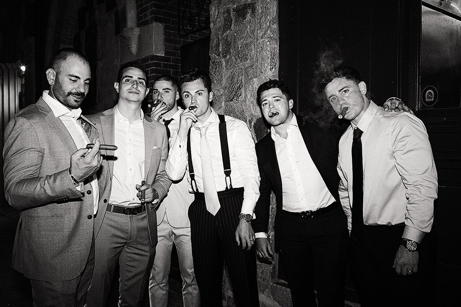 Six men dressed in formal attire, including suits, dress shirts, ties, and suspenders, stand closely together against a stone and brick wall. Each man is smoking a cigar, with some holding the cigars near their mouths and others holding them in their hands. The photo is in black and white, and the men appear to be enjoying a social moment.