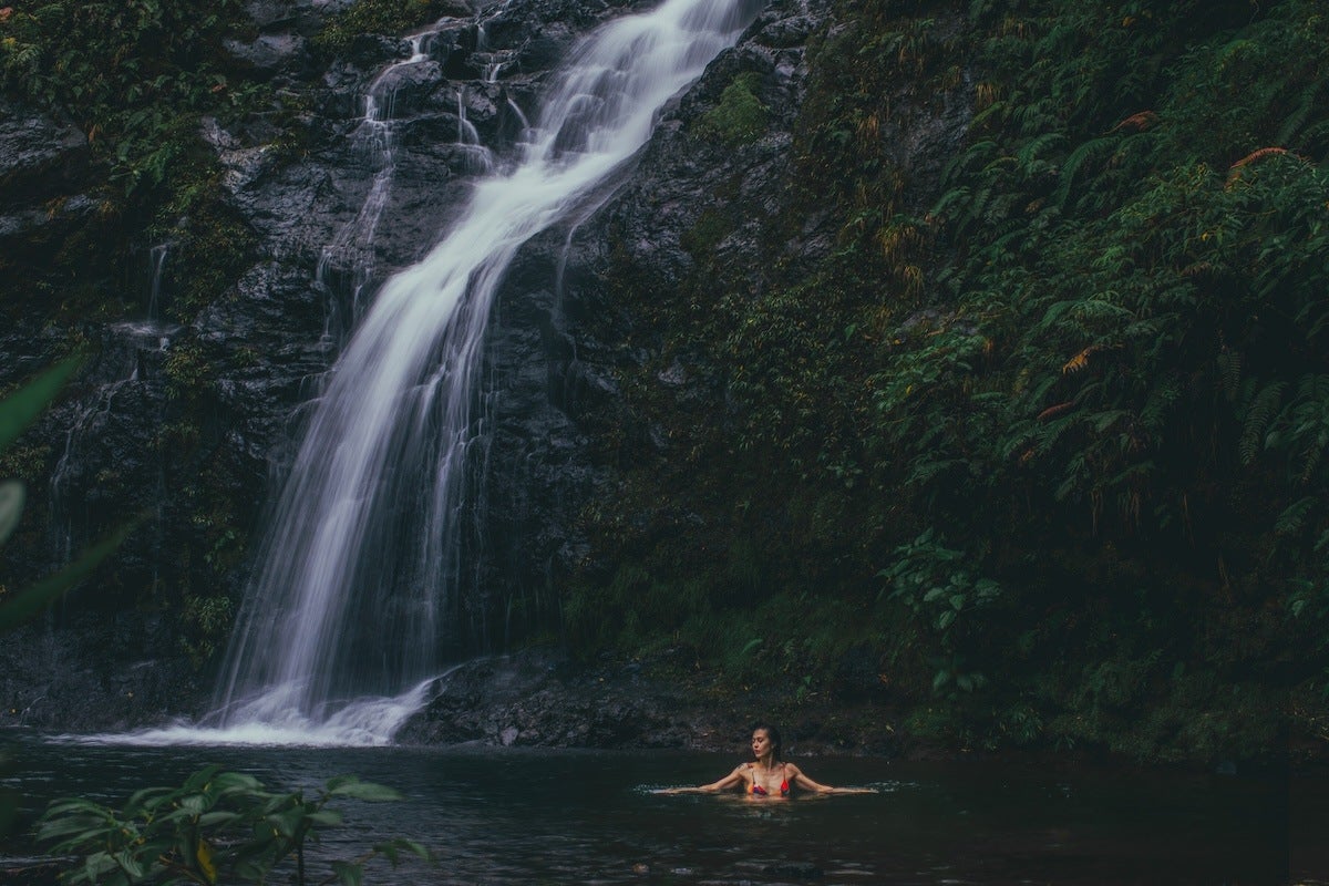 A woman in a red bikini is swimming in a dark natural pool at the base of a cascading waterfall surrounded by lush green foliage and rocky cliffs.