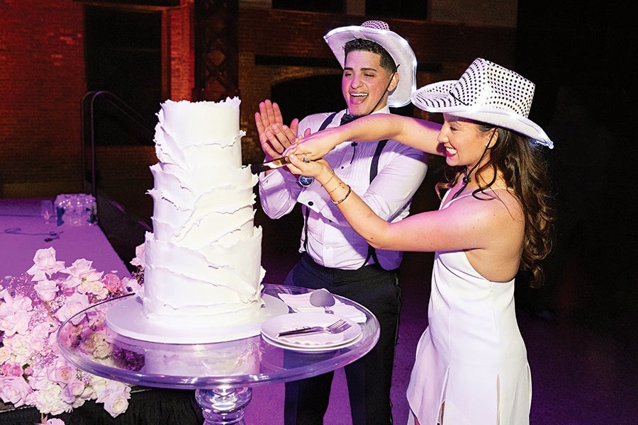 A couple wearing white cowboy hats is cutting a tall, white, textured wedding cake on a clear glass cake stand. The woman is in a white dress, and the man is in a white shirt with black suspenders and a bow tie. There are pink flowers on a table nearby.