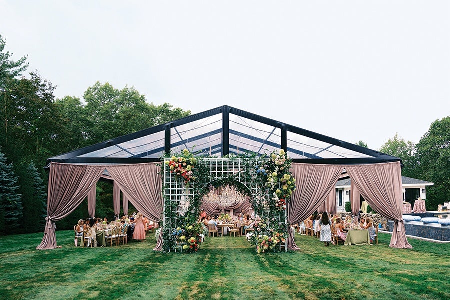 A large outdoor event tent with a transparent roof and mauve draped curtains tied back at the corners. The entrance features a white lattice arch decorated with green foliage and colorful flowers. Inside, guests are seated at round tables covered with beige tablecloths, and a large chandelier hangs from the center of the tent. The setting is on a grassy lawn surrounded by trees.
