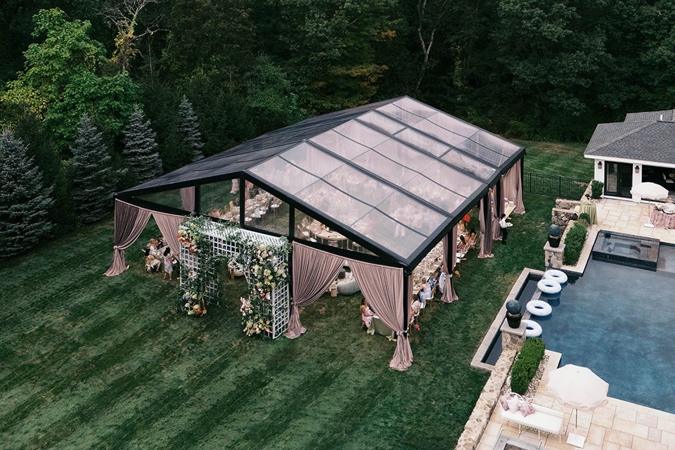 A large outdoor event tent with a transparent roof and mauve curtains is set up on a grassy lawn near a swimming pool. The tent features a floral archway at the entrance, and people are seated inside at tables. The surrounding area includes tall evergreen trees and a stone patio around the pool.
