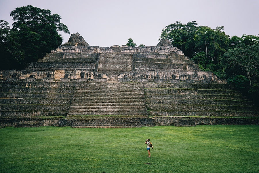 Large ancient stone pyramid with wide steps, surrounded by dense green trees, and a person standing on the grassy area in front of it.
