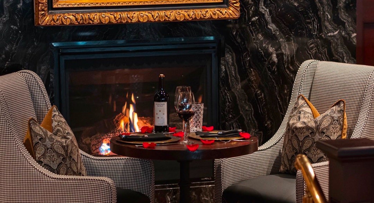 A cozy dining setup with a small round wooden table between two upholstered armchairs featuring a black and white houndstooth pattern. The table is set with two plates, black napkins, cutlery, a bottle of red wine, a wine glass, a water glass, and scattered red rose petals. In the background, there is a lit fireplace with a dark marble surround and a gold-framed painting above it. Each chair has two decorative pillows with intricate patterns in neutral tones and mustard yellow accents.