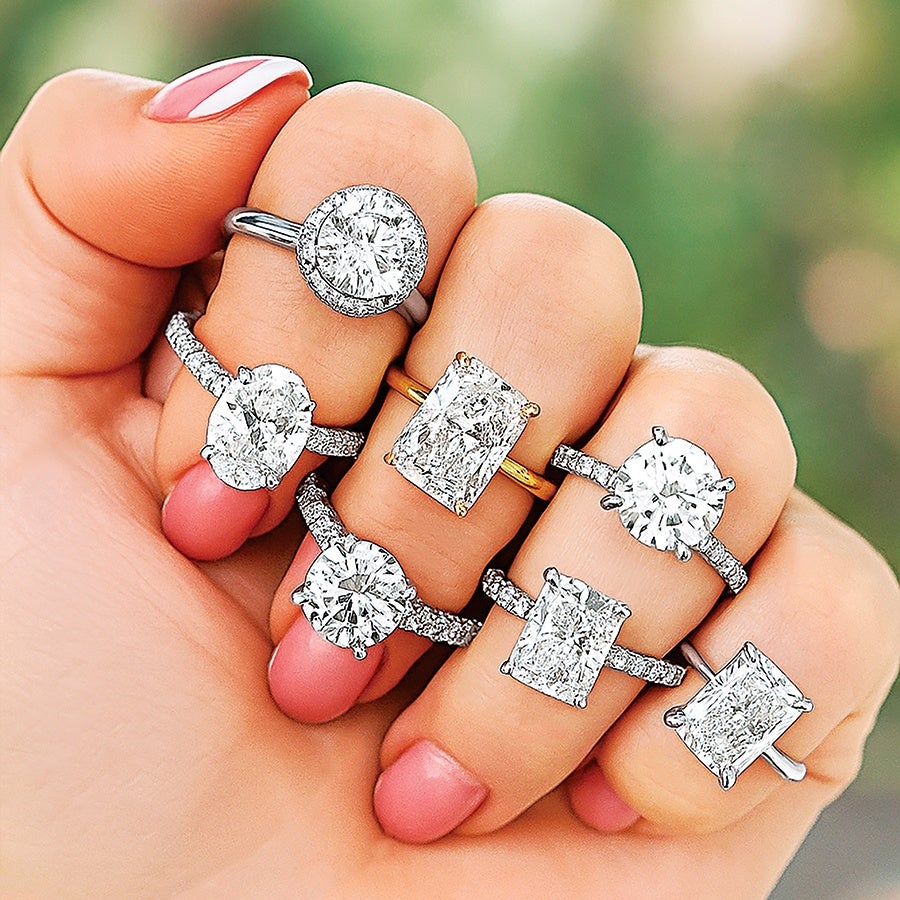 A hand wearing seven diamond rings, each with a different cut and setting. The rings feature round, oval, and rectangular diamonds set in silver or gold bands, with some bands adorned with smaller diamonds. The nails are painted pink with one nail having a white stripe. The background is blurred greenery.