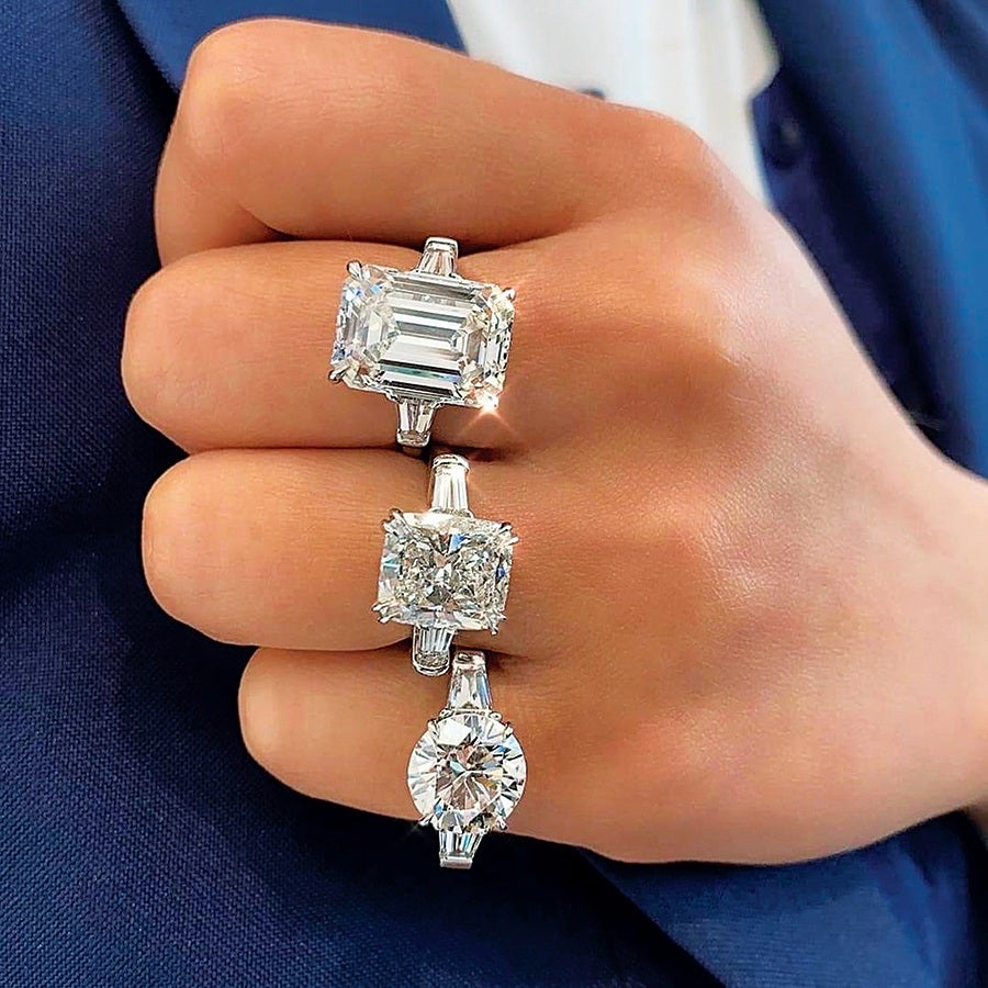 Three diamond rings with different cuts are worn on a hand. The top ring features an emerald-cut diamond, the middle ring has a cushion-cut diamond, and the bottom ring showcases a round brilliant-cut diamond. Each ring is set in a silver or white gold band with baguette side stones. The hand is positioned against a dark blue fabric background.