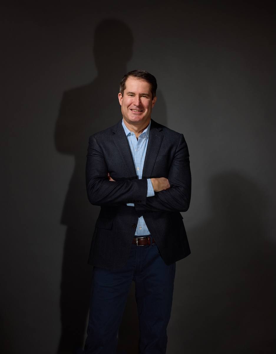 Man standing with arms crossed, wearing a dark blazer over a light blue checkered shirt and dark pants, smiling against a dark background with his shadow visible behind him.