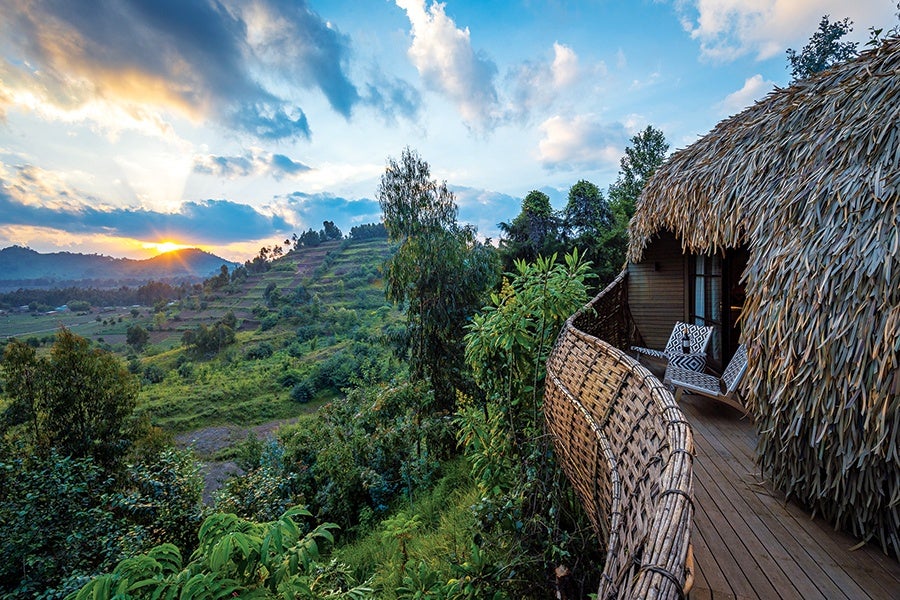 A wooden balcony with woven railing and two patterned chairs extends from a thatched-roof hut, overlooking lush green hills and trees under a partly cloudy sky at sunset.