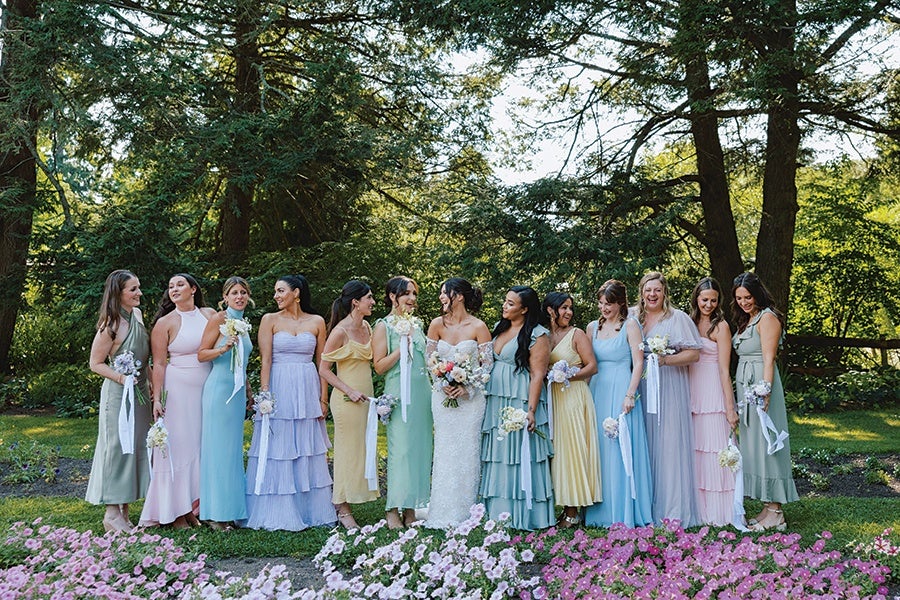 A group of twelve women standing outdoors in a garden setting, dressed in pastel-colored formal dresses. The woman in the center is wearing a white strapless wedding gown and holding a bouquet of flowers, while the others wear dresses in shades of green, blue, pink, yellow, and lavender, each holding small bouquets. The background features tall trees and greenery, with pink and white flowers in the foreground.