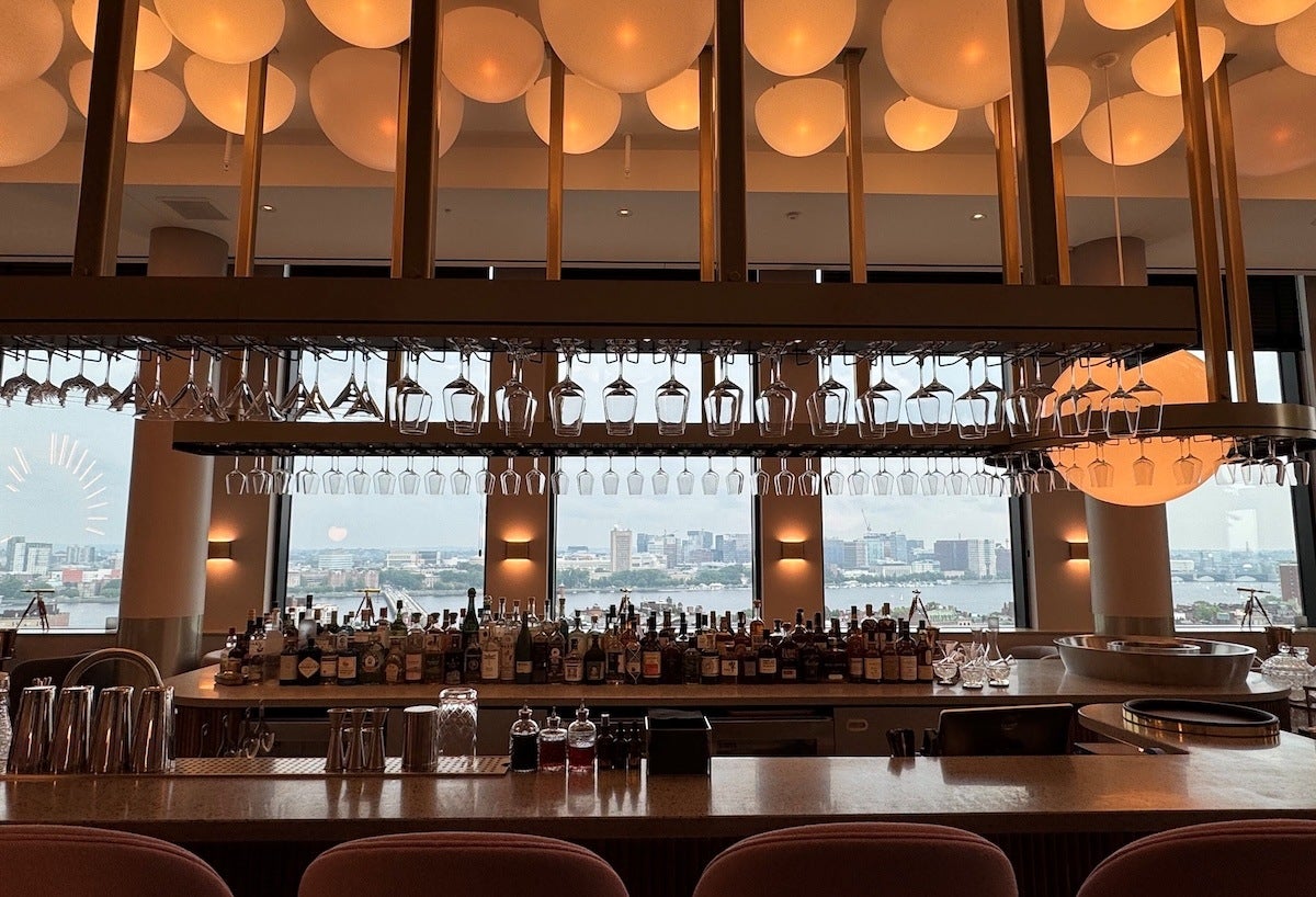 Modern bar with a wide selection of liquor bottles arranged on the counter, multiple rows of hanging wine glasses above, and large windows in the background offering a cityscape view. The ceiling features round, warm-toned light fixtures, and the bar counter has various bar tools and glassware.
