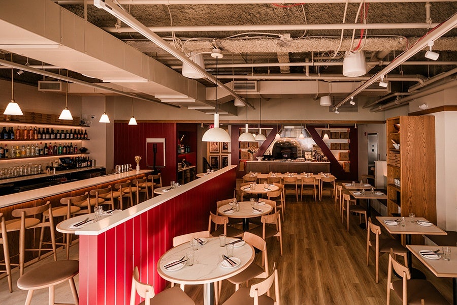A modern restaurant interior featuring wooden floors and light wood chairs paired with round and square marble-top tables. A red-paneled bar with high stools runs along the left side, stocked with various bottles and glasses. Pendant lights hang from the ceiling, illuminating the space. The back wall has a wood-fired oven surrounded by shelves holding pizza boxes and framed pictures. The ceiling exposes ductwork and pipes, adding an industrial touch.