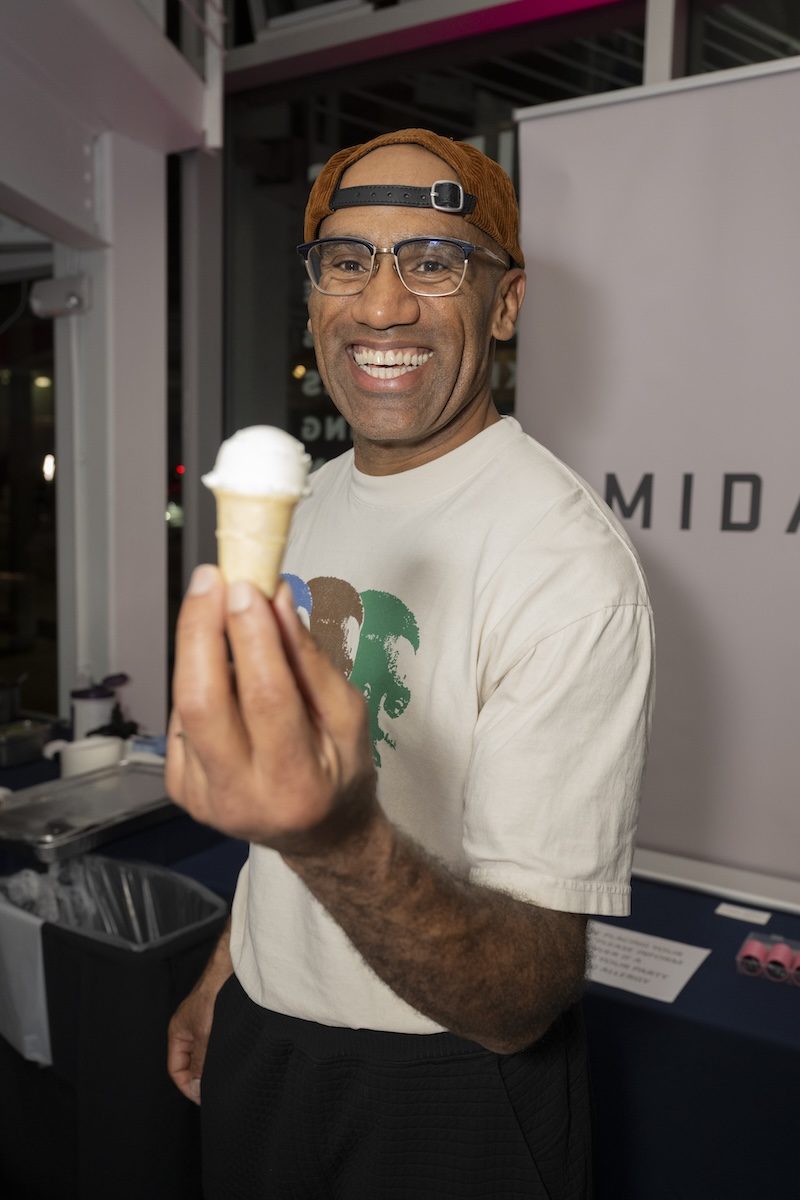 A smiling man wearing glasses, a brown backward cap, and a beige t-shirt holds a small ice cream cone with a scoop of white ice cream toward the camera.