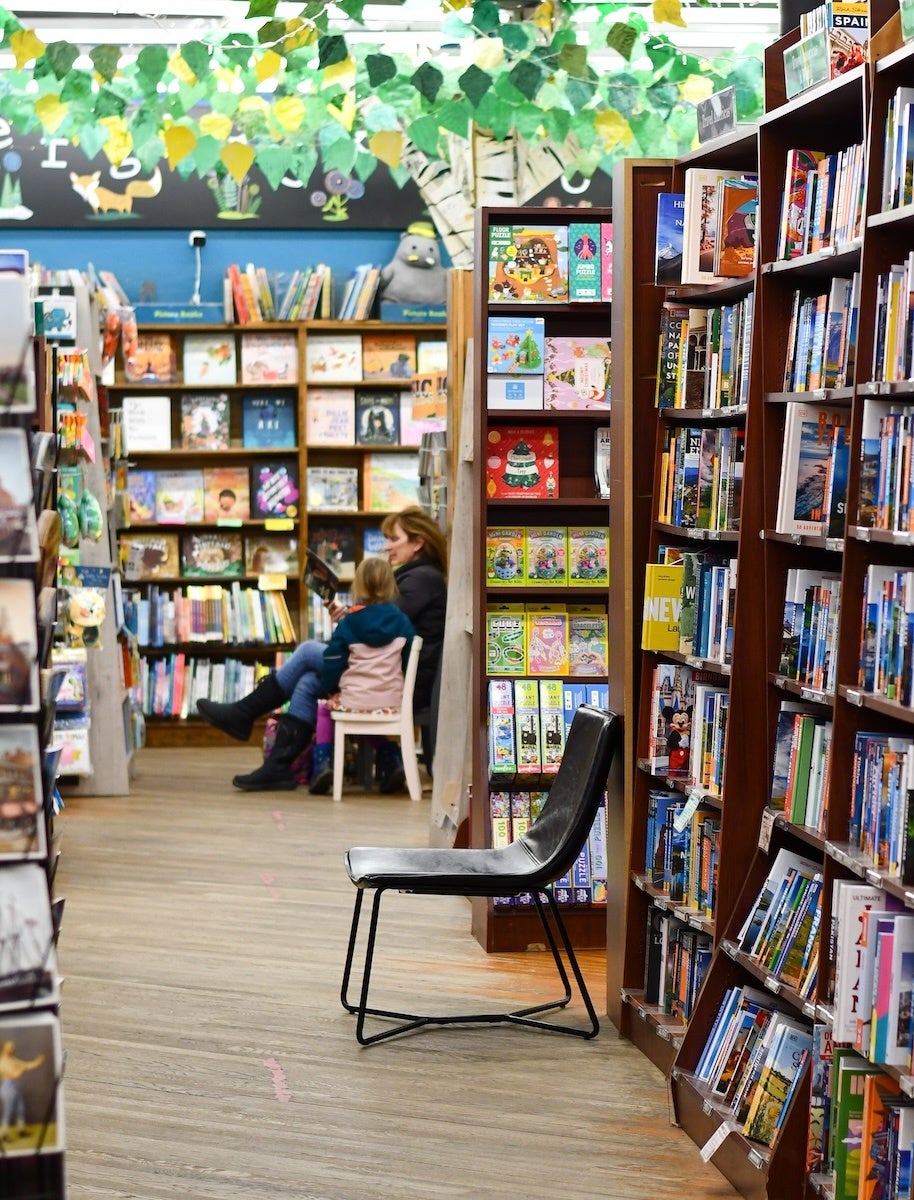 A black chair with a metal frame is positioned in the aisle of a bookstore. The bookstore has wooden floors and tall bookshelves filled with colorful books on both sides. In the background, a woman and a child are seated, with the woman reading a book to the child. The ceiling is decorated with green and yellow leaf-shaped decorations, and a mural featuring a fox and flowers is visible on the back wall.