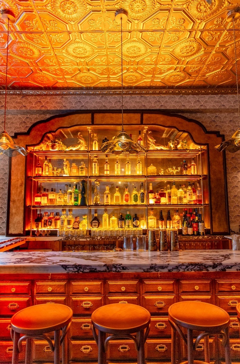 A vintage-style bar with a marble countertop and wooden drawers beneath. Behind the bar is a backlit shelf filled with various bottles of liquor, arranged neatly. The ceiling features ornate, gold-toned embossed tiles, and three pendant lights with decorative shades hang above the bar. Three wooden bar stools with round, cushioned seats are positioned in front of the counter. The overall ambiance is warm and inviting with a classic, elegant design.
