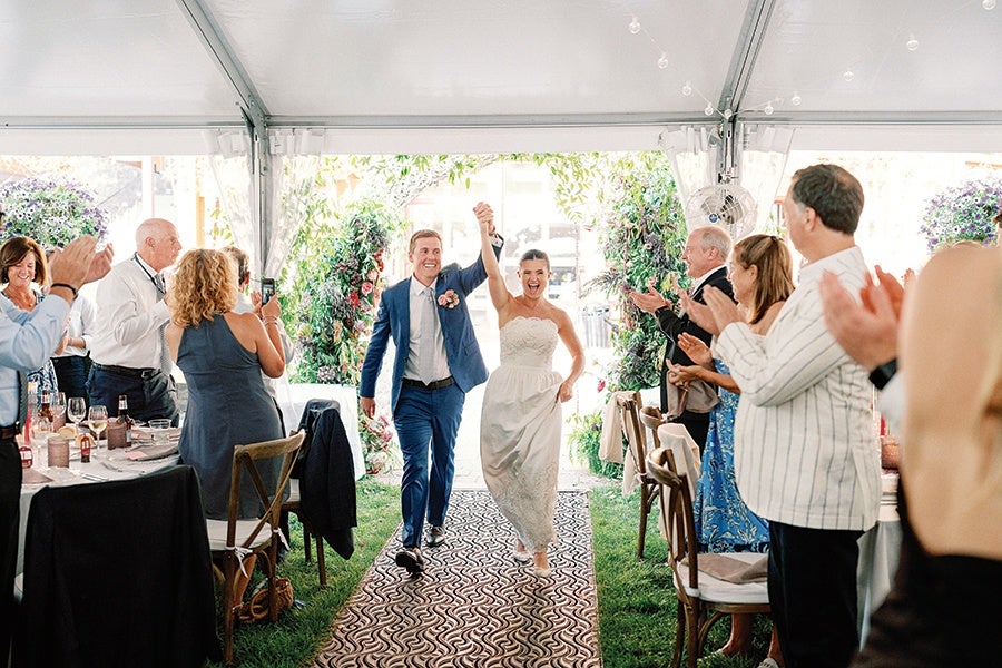 A bride and groom walk hand in hand down an aisle covered with a patterned carpet inside a white tent. The bride wears a strapless white wedding dress and the groom is dressed in a blue suit with a white shirt. Guests on both sides of the aisle stand and applaud, some taking photos, with greenery and floral decorations visible in the background.