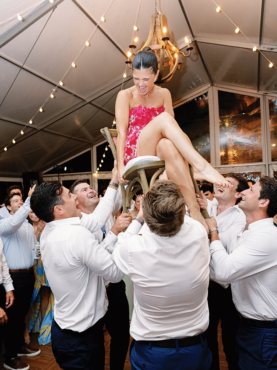 A woman in a strapless red and white dress is being lifted on a wooden chair by five men dressed in white shirts and dark pants. The woman is smiling and laughing, with her hair tied up. The scene takes place indoors under a tent with string lights and a chandelier overhead. Several people in the background are watching and smiling.