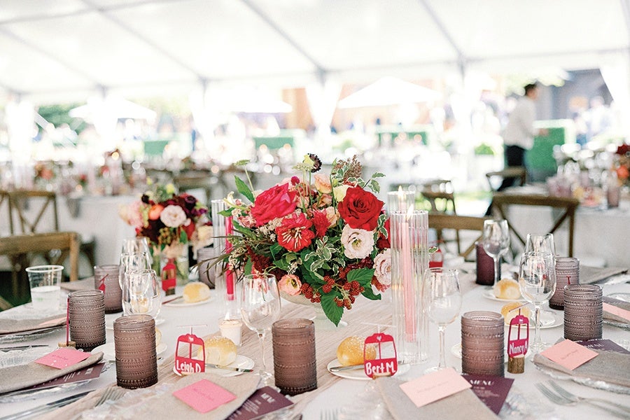 A round table set for an event with a centerpiece of red and pink flowers and greenery. The table has multiple wine glasses, water glasses, and brown textured candle holders. There are place cards and small name tags with red backgrounds and white text, along with beige napkins and plates with bread rolls. The background shows other similarly set tables under a white tent.