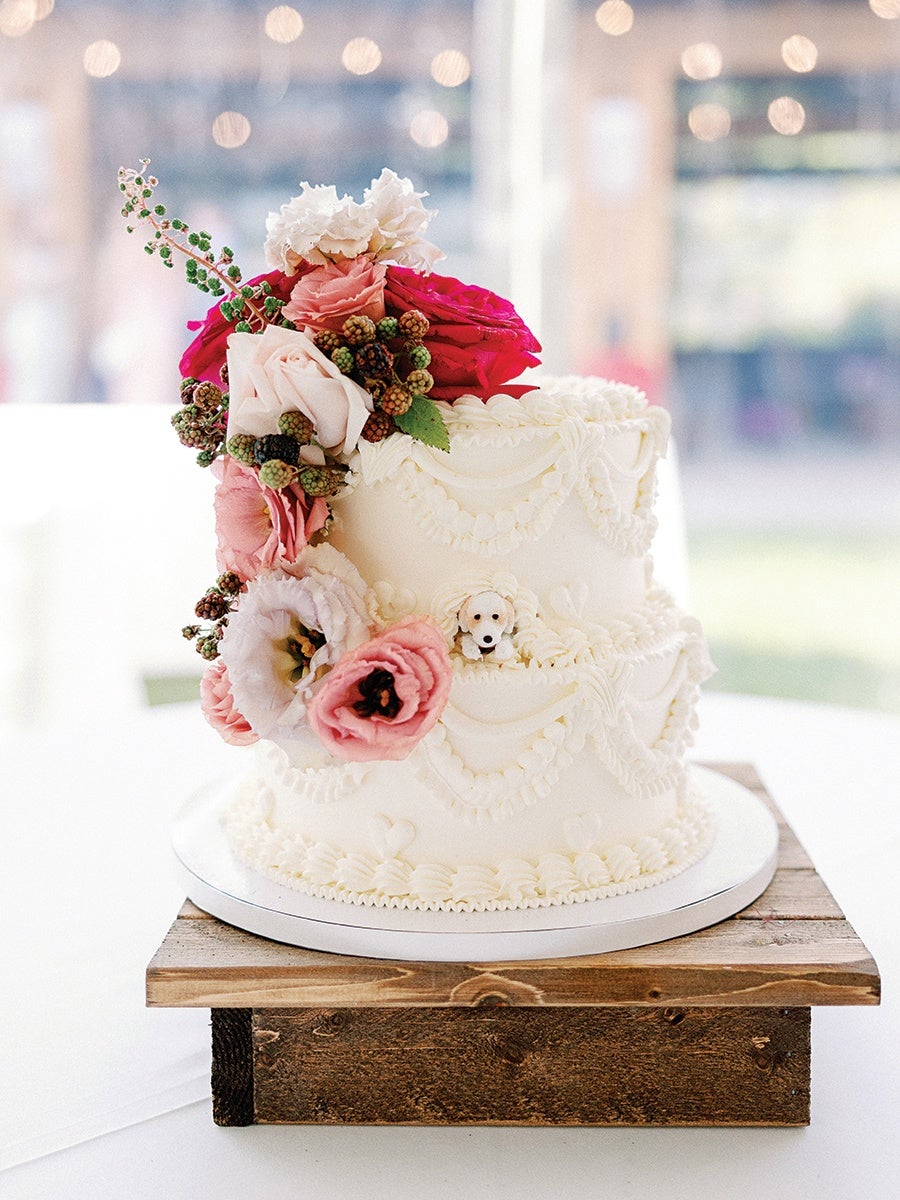 Two-tier white cake decorated with intricate white icing patterns, topped and adorned on one side with a cluster of pink, red, and white flowers and green berries, placed on a round white cake board and a rustic wooden stand.