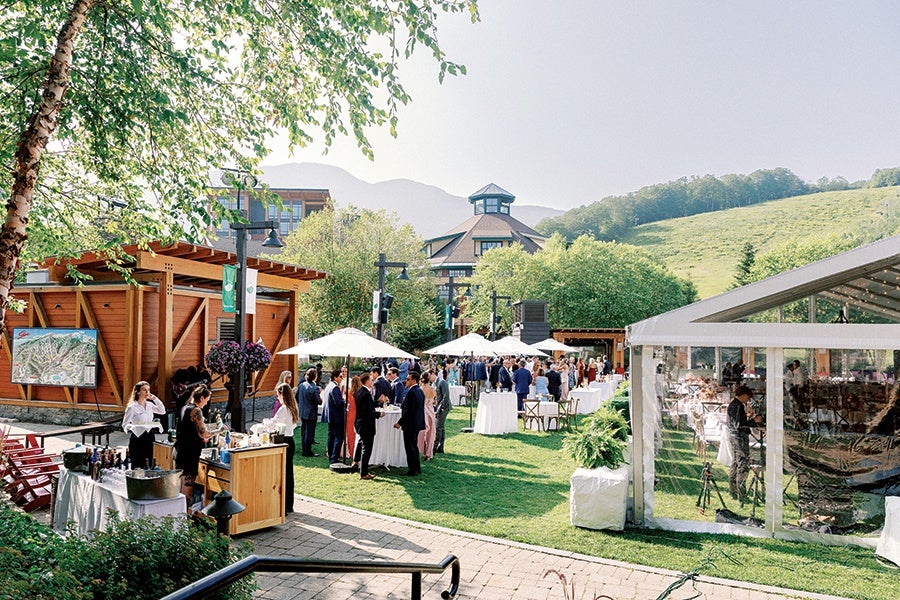 Outdoor event with people dressed in formal attire gathered around cocktail tables with white umbrellas on a grassy area. A wooden building and a large tent with transparent sides are visible, along with trees and a hillside in the background.