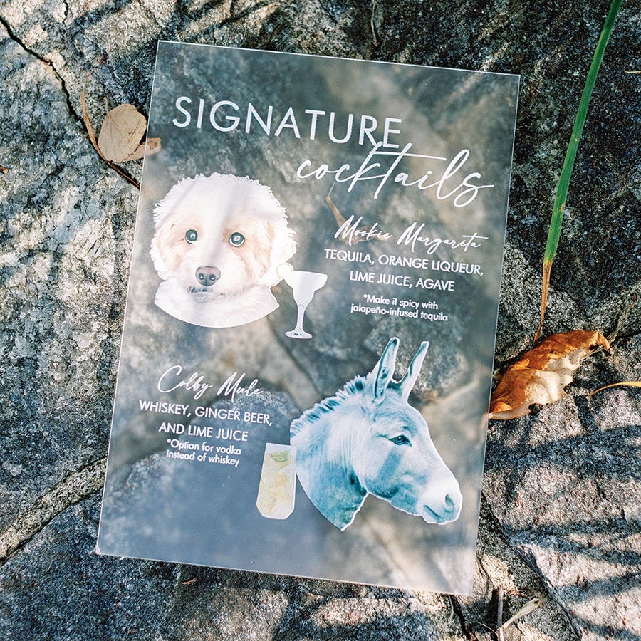 A clear acrylic sign on a stone surface displaying "Signature cocktails" with two illustrated animal heads: a white dog and a blue donkey. The dog represents the "Muttie Margarita" cocktail with ingredients tequila, orange liqueur, lime juice, and agave, with a note to make it spicy with jalapeño-infused tequila. The donkey represents the "Colby Mule" cocktail with whiskey, ginger beer, and lime juice, with an option for vodka instead of whiskey. Each cocktail is accompanied by a small illustration of its respective drink.