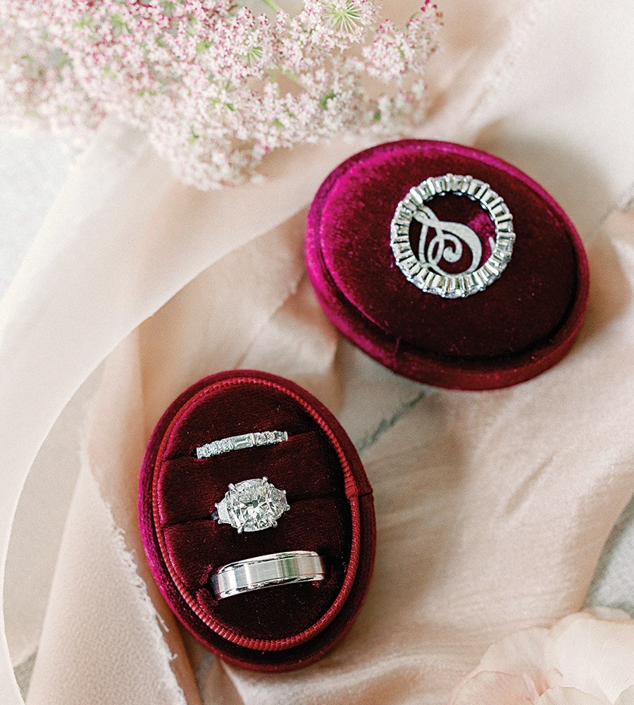 Three silver rings with diamonds are displayed in a red velvet oval ring box. The box is open, showing a delicate floral arrangement and soft beige fabric in the background.