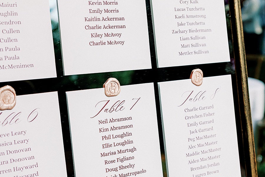 A seating chart with multiple white cards displaying table numbers and guest names in elegant script and serif fonts. Each card is attached to a reflective surface with a small, round, beige wax seal featuring an embossed monogram. The visible table numbers include 6, 7, and 8, with names listed below each. The background is slightly blurred, emphasizing the seating cards.