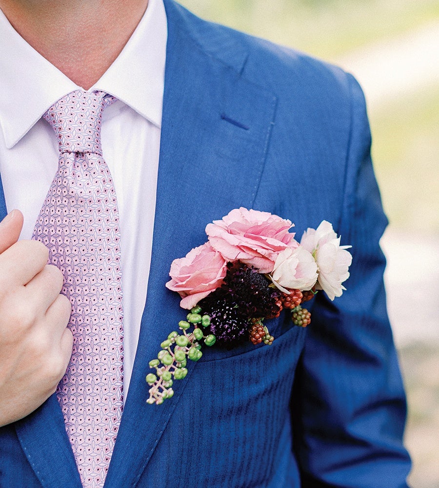 A person wearing a blue suit jacket with a white dress shirt and a light pink patterned tie. The suit jacket has a boutonniere pinned to the left lapel pocket, featuring pink and white flowers along with small green and dark purple berries. The person is holding the tie with their left hand.