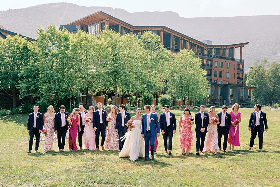 A wedding party walking on a grassy lawn in front of a large building with trees and mountains in the background. The bride is wearing a white gown and holding a bouquet, while the groom is in a blue suit with a pink tie. Bridesmaids are dressed in various shades of pink and floral dresses, and groomsmen are in dark suits with pink ties. The group appears joyful and relaxed.