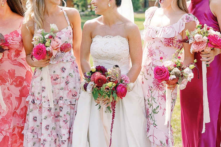 A bride in a strapless white lace wedding gown holding a vibrant bouquet of mixed flowers including pink and red blooms, surrounded by bridesmaids wearing floral and solid pink dresses, each holding similar colorful bouquets.