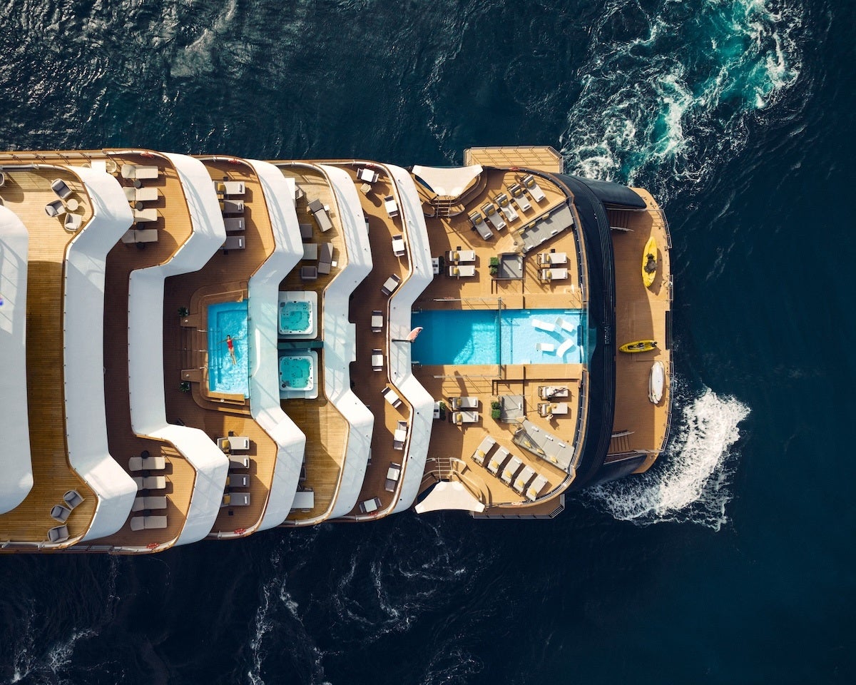 Aerial view of the rear deck of a luxury cruise ship featuring multiple levels with white awnings, sun loungers, two hot tubs, a large swimming pool, and small boats including kayaks on the side. The ship is sailing on dark blue ocean water.