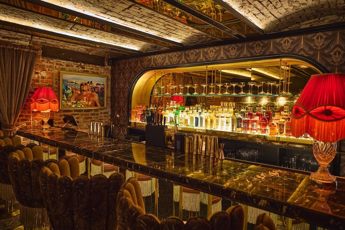 Luxurious bar interior with a polished dark marble countertop, ornate velvet bar stools with fringe detailing, and warm ambient lighting. The back wall features a large mirror with shelves holding various bottles of liquor and upside-down cocktail glasses. Two vintage-style red lamps with fringed shades add a cozy glow, and exposed brick walls and patterned wallpaper contribute to the rich, elegant atmosphere. A framed photograph hangs on the brick wall beside heavy curtains.