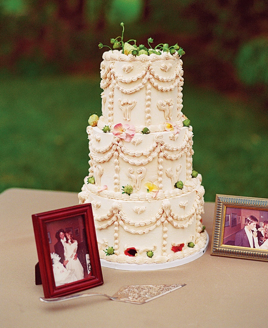 Three-tiered cream-colored cake decorated with intricate piped icing swags and floral patterns, topped with small green berries and a few pastel flower petals. The cake is placed on a beige tablecloth, flanked by two framed vintage wedding photos and a silver cake server.