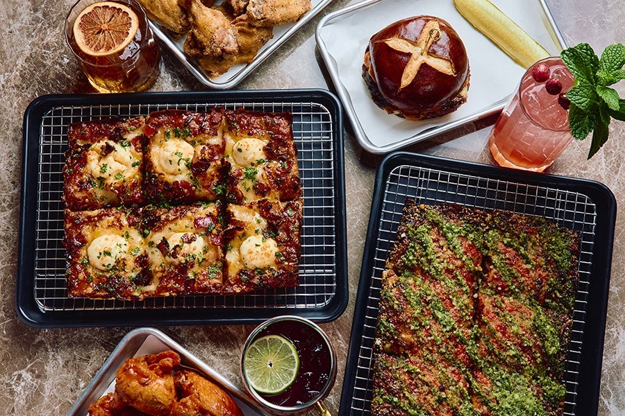 Two rectangular trays of Detroit-style pizza, one topped with melted cheese and herbs, the other with a green herb sauce and red seasoning. A tray with fried chicken wings, a sandwich with a pretzel bun and a pickle spear, and two drinks—one garnished with a dried citrus slice and the other with fresh mint and cranberries—are also visible on a marble surface.