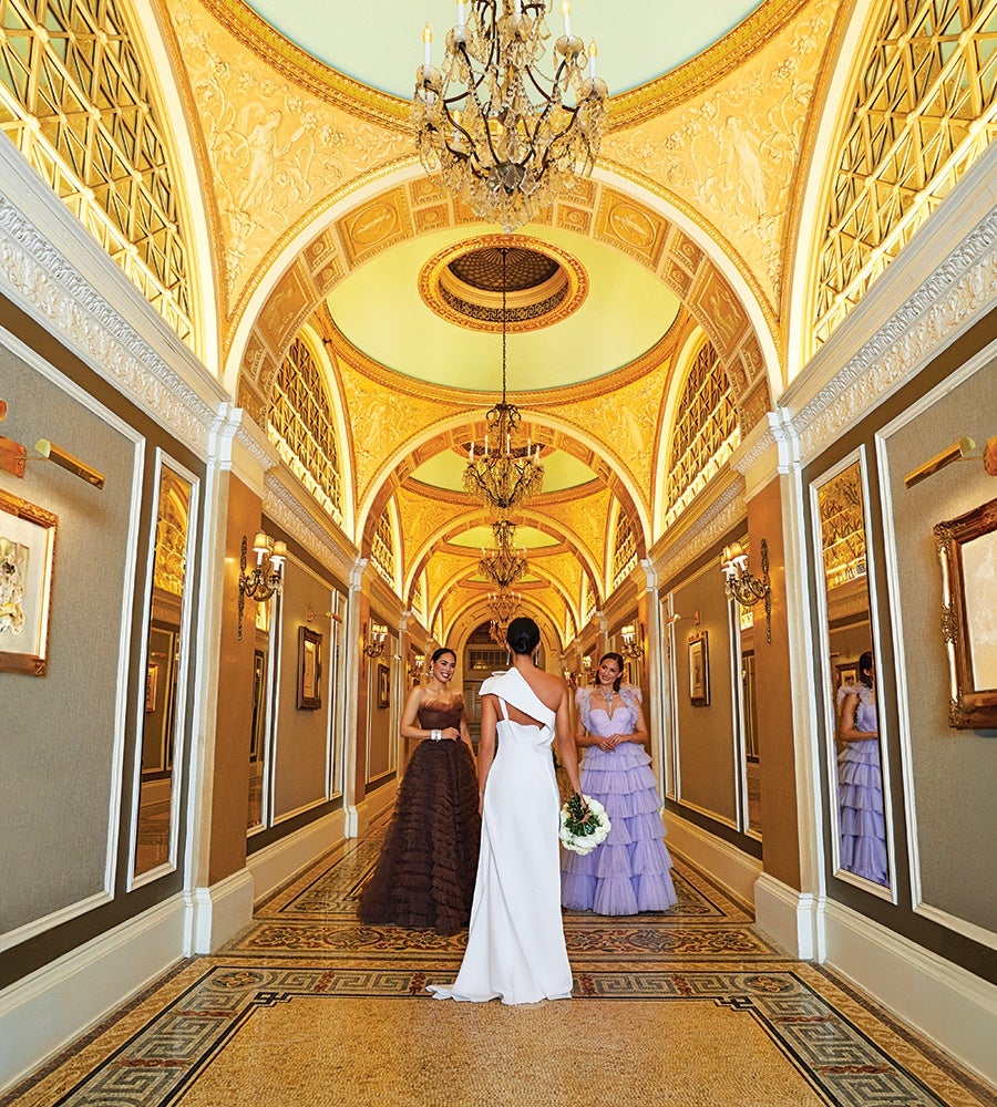 A woman in a white, one-shoulder gown holding a bouquet stands in an ornate hallway with a golden arched ceiling and chandeliers. She faces two women in elegant dresses, one in a dark brown strapless gown and the other in a lavender tiered dress. The hallway features large mirrors, framed artwork, and intricate tile flooring.