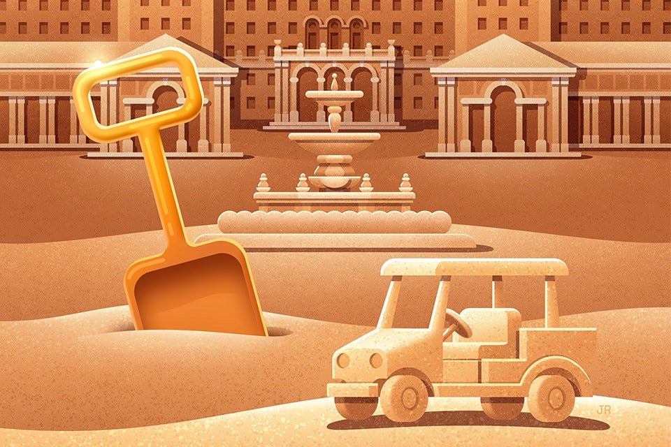 A large orange shovel partially buried in sand next to a small sand sculpture of a car, set against a background of sand sculptures resembling classical buildings and a fountain.
