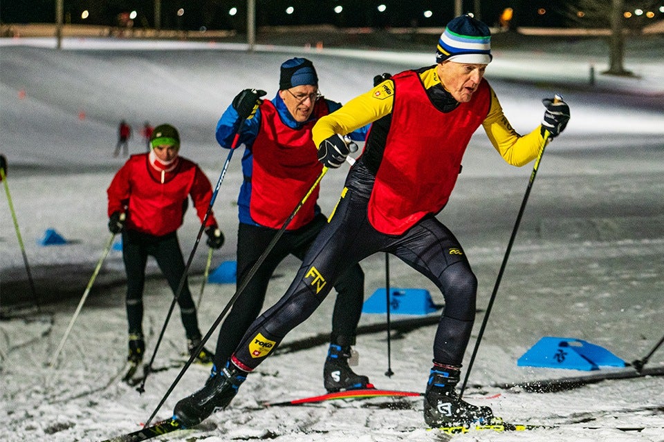 Three cross-country skiers wearing red bibs are skiing on a snowy trail at night. The skier in the foreground is dressed in a yellow and black outfit with a striped beanie, while the two skiers behind wear darker clothing and hats. Blue markers are visible on the snow along the trail.