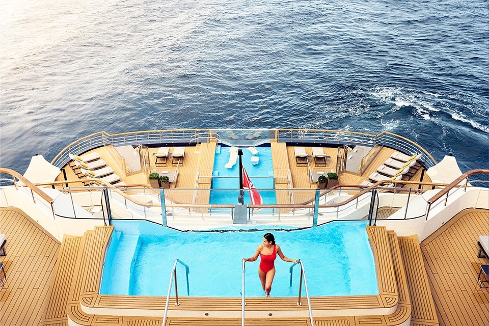 A woman in a red swimsuit is stepping into a rectangular pool on the deck of a yacht. The yacht deck features wooden flooring, lounge chairs, tables, and a smaller pool area with sunbeds and a flag in the background, surrounded by the ocean.