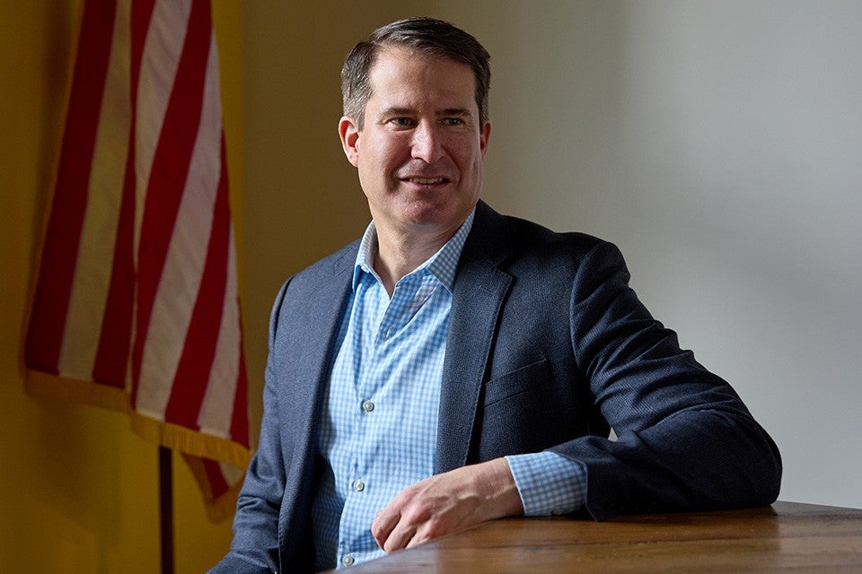 A man wearing a blue checkered shirt and a dark blazer is sitting at a wooden table. An American flag is visible in the background on the left side. The man is looking slightly to his right with a slight smile.