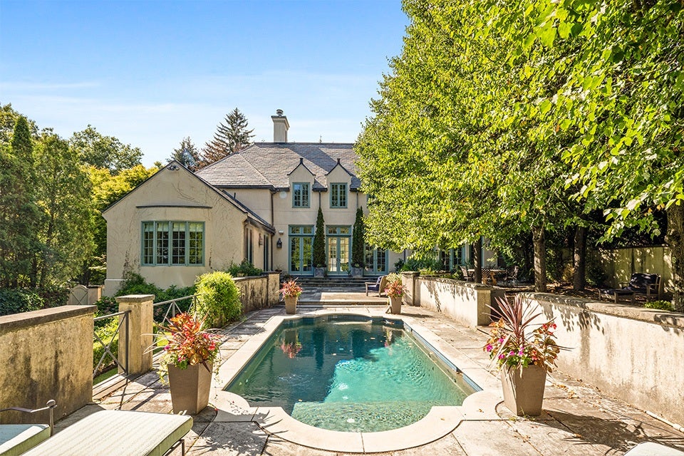 A large, elegant house with beige exterior walls and green-framed windows is set behind a rectangular swimming pool. The pool is surrounded by a stone patio with four large planters filled with colorful flowers. On the right side, tall leafy trees provide shade, while the left side features a low stone wall and lush greenery. The sky is clear and blue, enhancing the bright and inviting atmosphere.