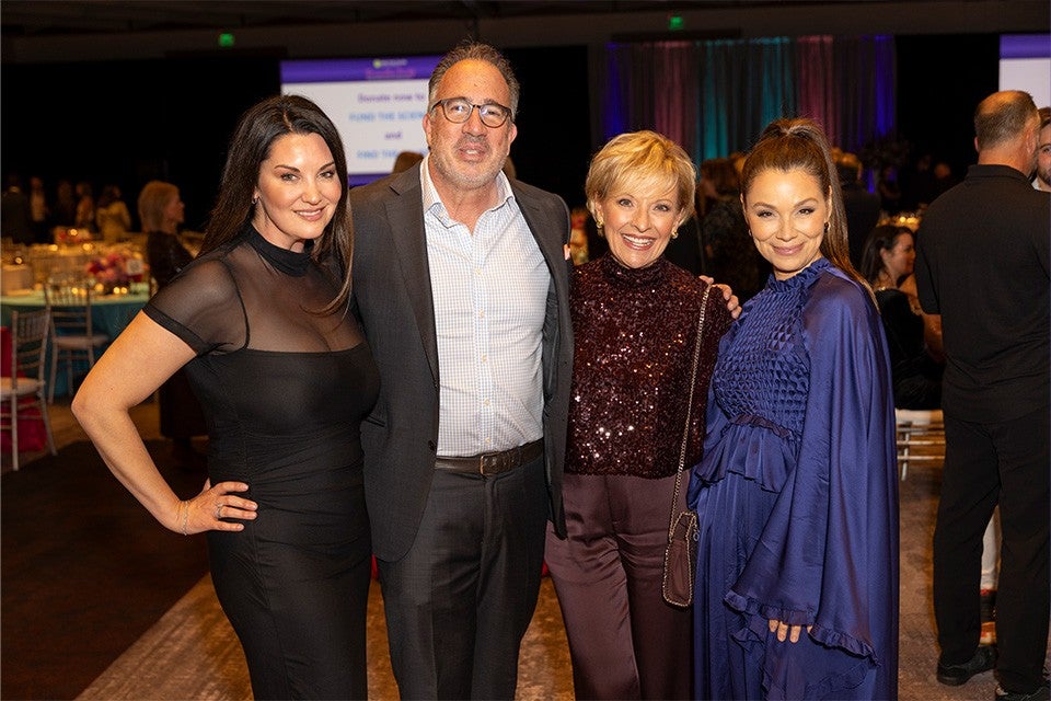 Four people stand closely together indoors at an event. From left to right: a woman with long dark hair wearing a fitted black dress with sheer sleeves; a man with glasses, short hair, and a beard wearing a dark suit and light checkered shirt; a woman with short blonde hair wearing a dark sequined top and matching pants, holding a small purse; and a woman with long hair tied back wearing a flowing dark blue dress with textured detailing. The background shows other attendees and tables with chairs.