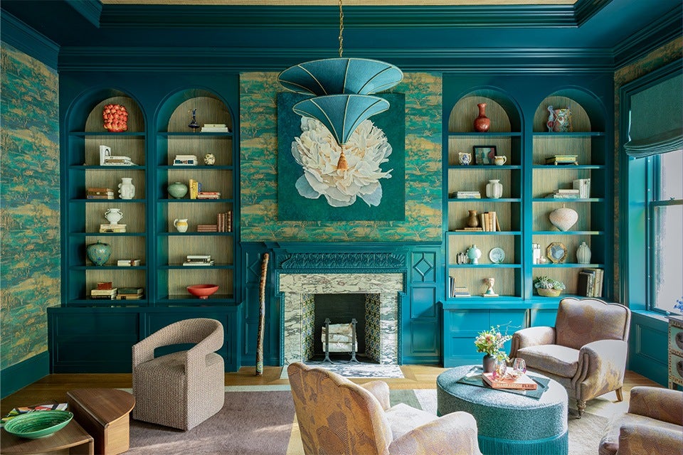 A teal-themed living room with built-in arched bookshelves flanking a marble fireplace. The shelves hold various decorative items and books. Above the fireplace is a large floral artwork. The room features a teal chandelier, patterned wallpaper, and a mix of beige and teal upholstered chairs around a round teal ottoman with a tray and flowers. A window with a teal frame and Roman shade is on the right side.