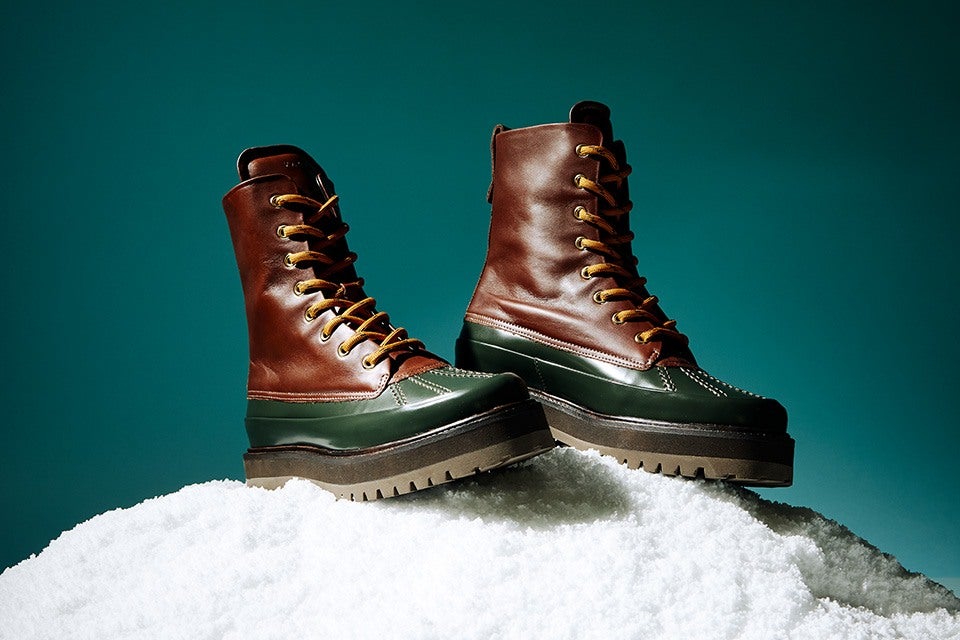 Pair of brown and dark green leather boots with yellow laces, thick rubber soles, displayed on a mound of white snow against a teal background.