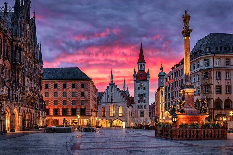 A historic European town square at sunset with a dramatic pink and purple sky. The square features ornate Gothic and Renaissance-style buildings, including a tall clock tower with a red pointed roof in the center. On the right, there is a tall column topped with a golden statue, surrounded by smaller statues and decorative lamps. The ground is paved with large stone tiles, and warm lights illuminate the buildings and statues, creating a cozy atmosphere.